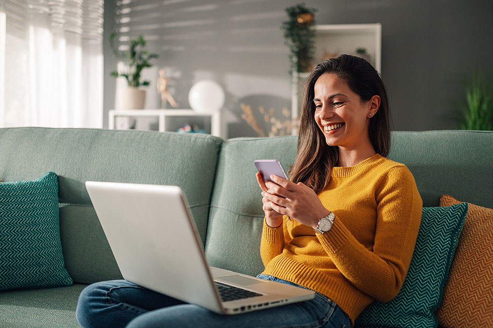 Woman sitting on a sofa at home, using a laptop and phone to research gummy vitamins.