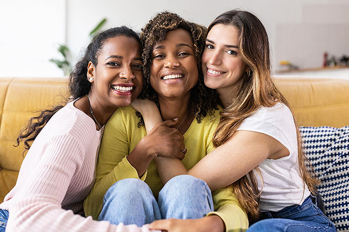 Three women sitting together on a couch, smiling and hugging each other.